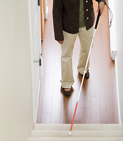 Blinde Frau mit Blindenstock vor Treppe.